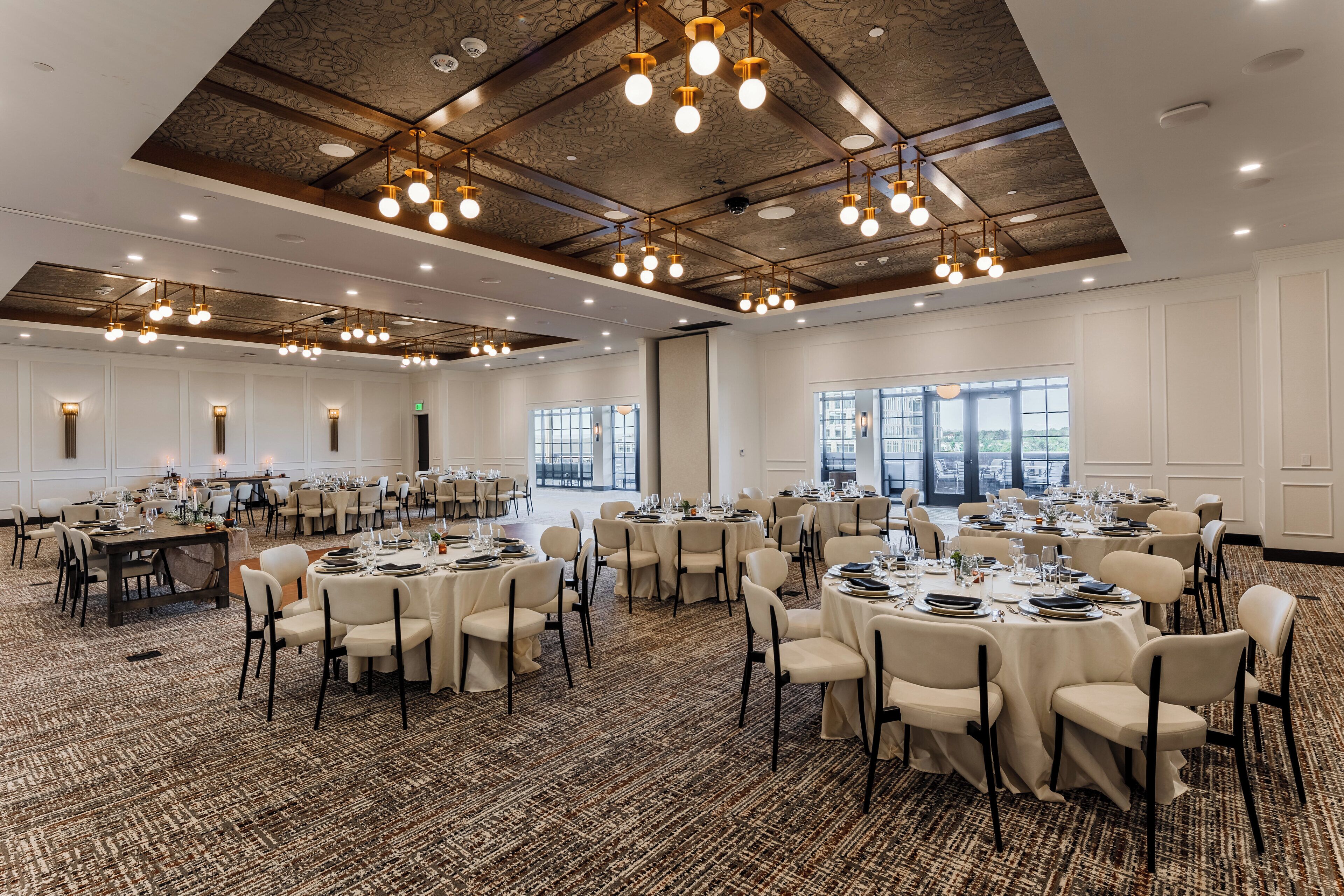 Un amplio salón de banquetes con mesas redondas dispuestas para el servicio de comidas, techos altos con vigas a la vista y grandes ventanales que dejan entrar luz natural.