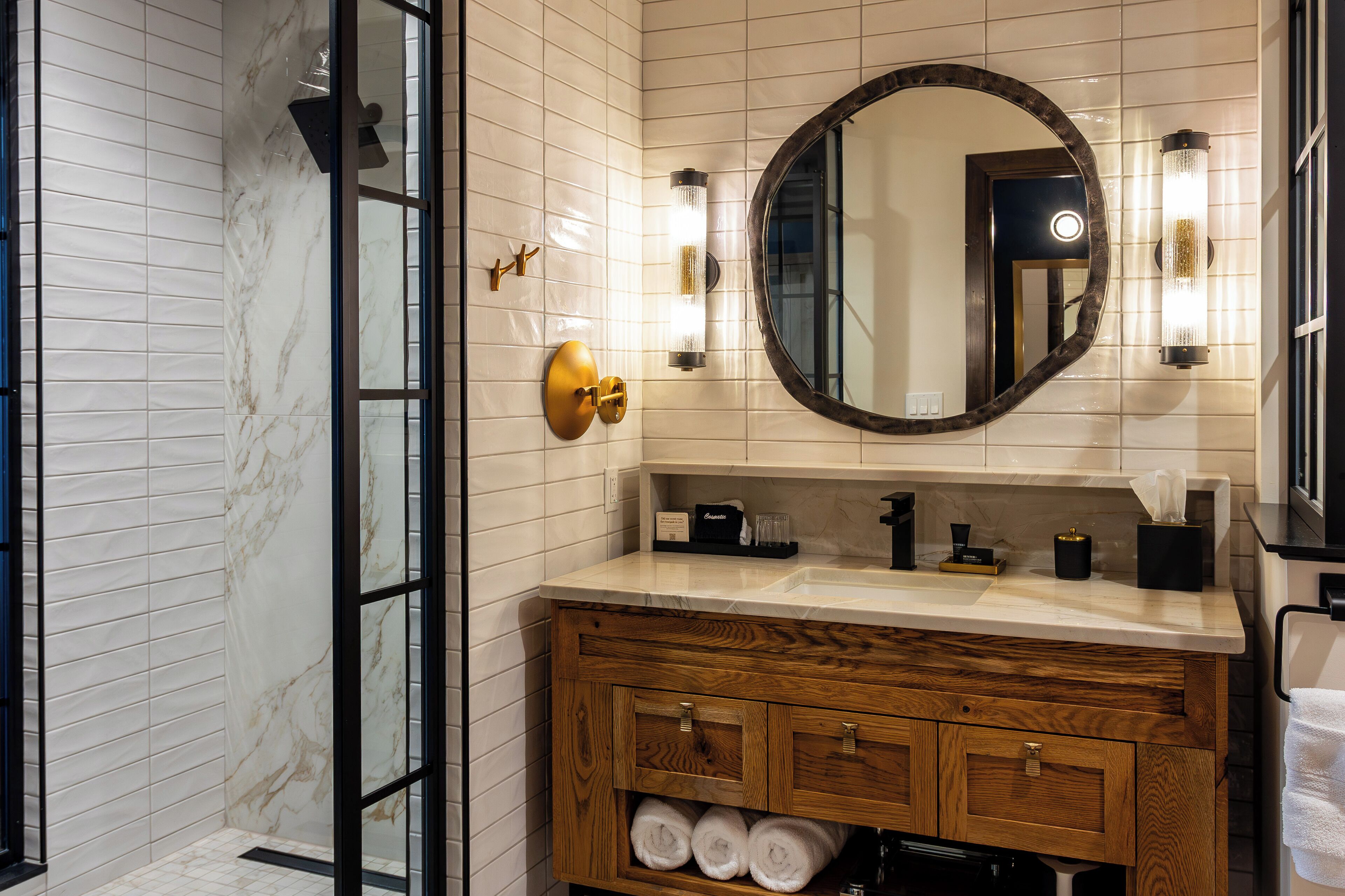 A bathroom with a wooden vanity, a round mirror, and a marble-tiled wall.