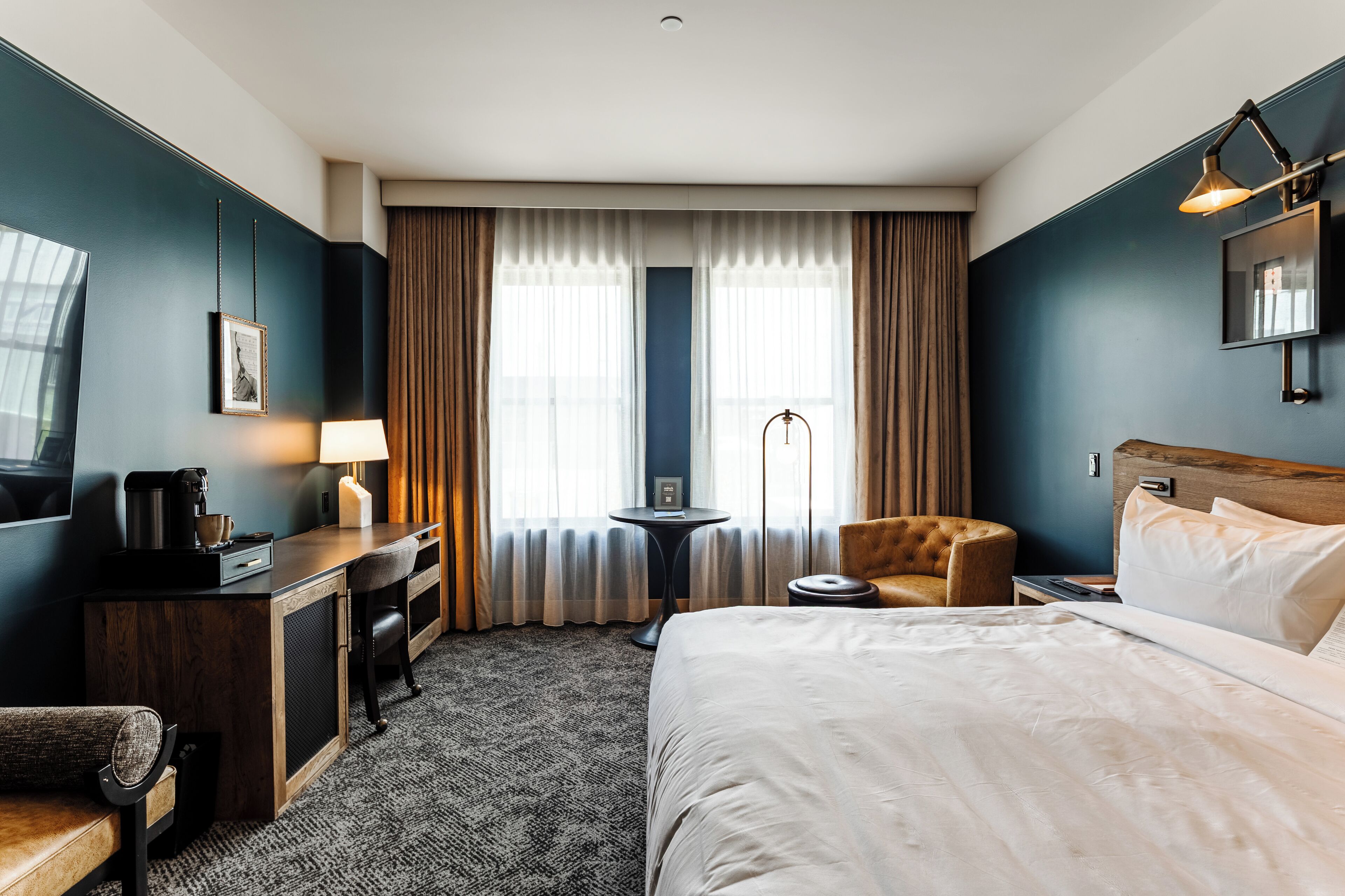 A hotel room with a bed, a desk with a lamp, a chair, a small table with a lamp, and a television.