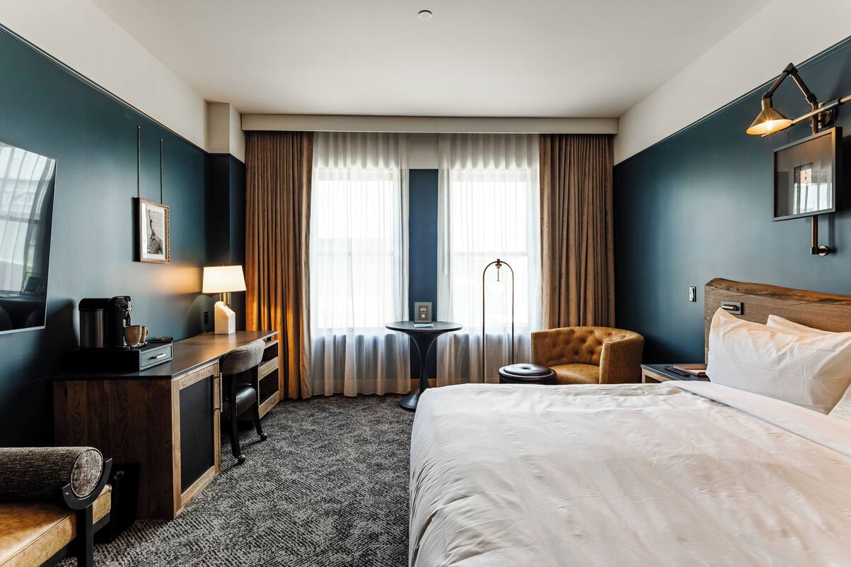 A hotel room with a bed, a desk with a lamp, a chair, a small table with a lamp, and a television.