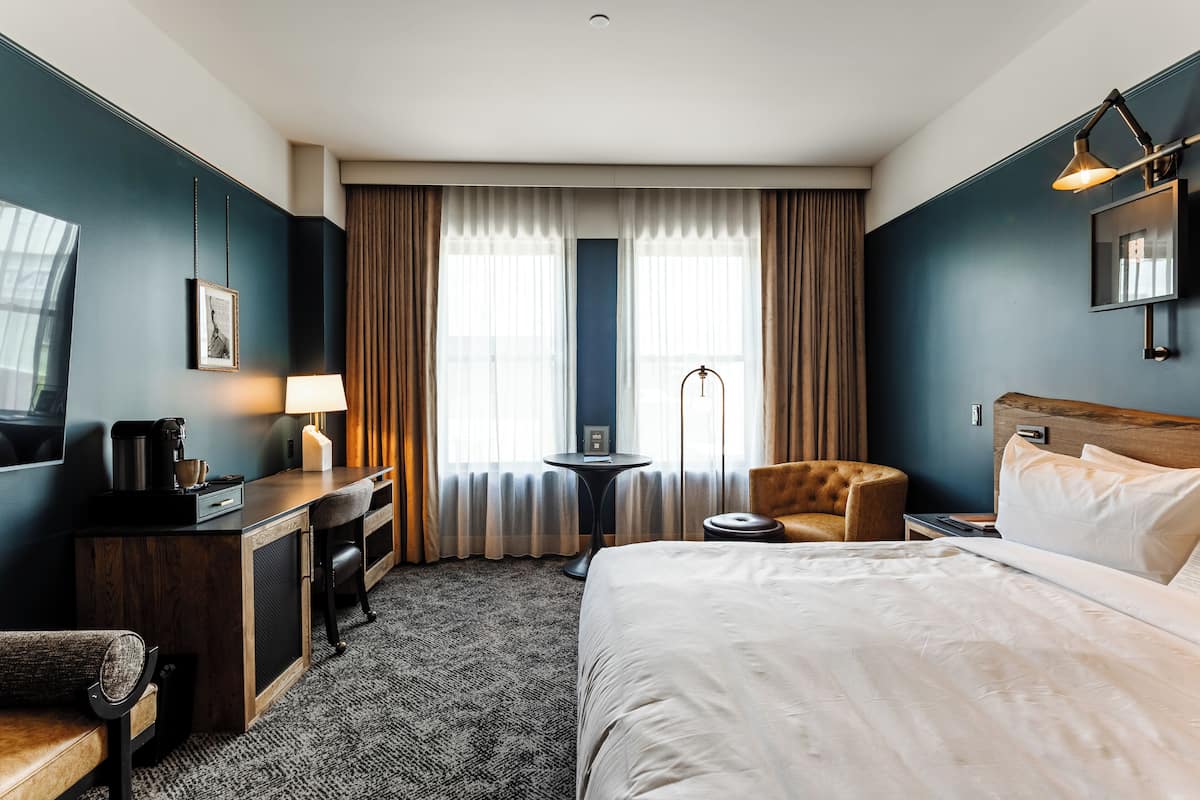 A hotel room with a bed, a desk with a lamp, a chair, a small table with a lamp, and a television.