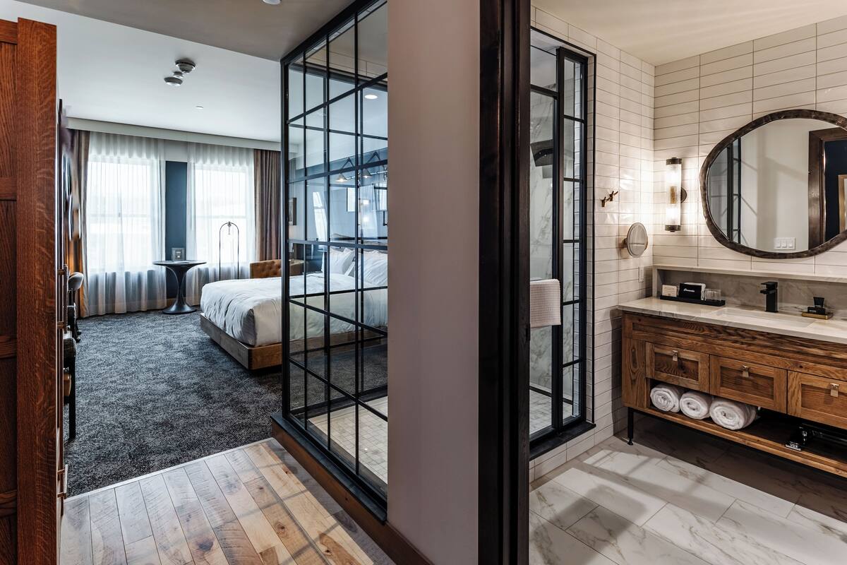 A modern hotel room with a large bed, a glass partition, a wooden vanity with a mirror, and a view of the city through large windows.