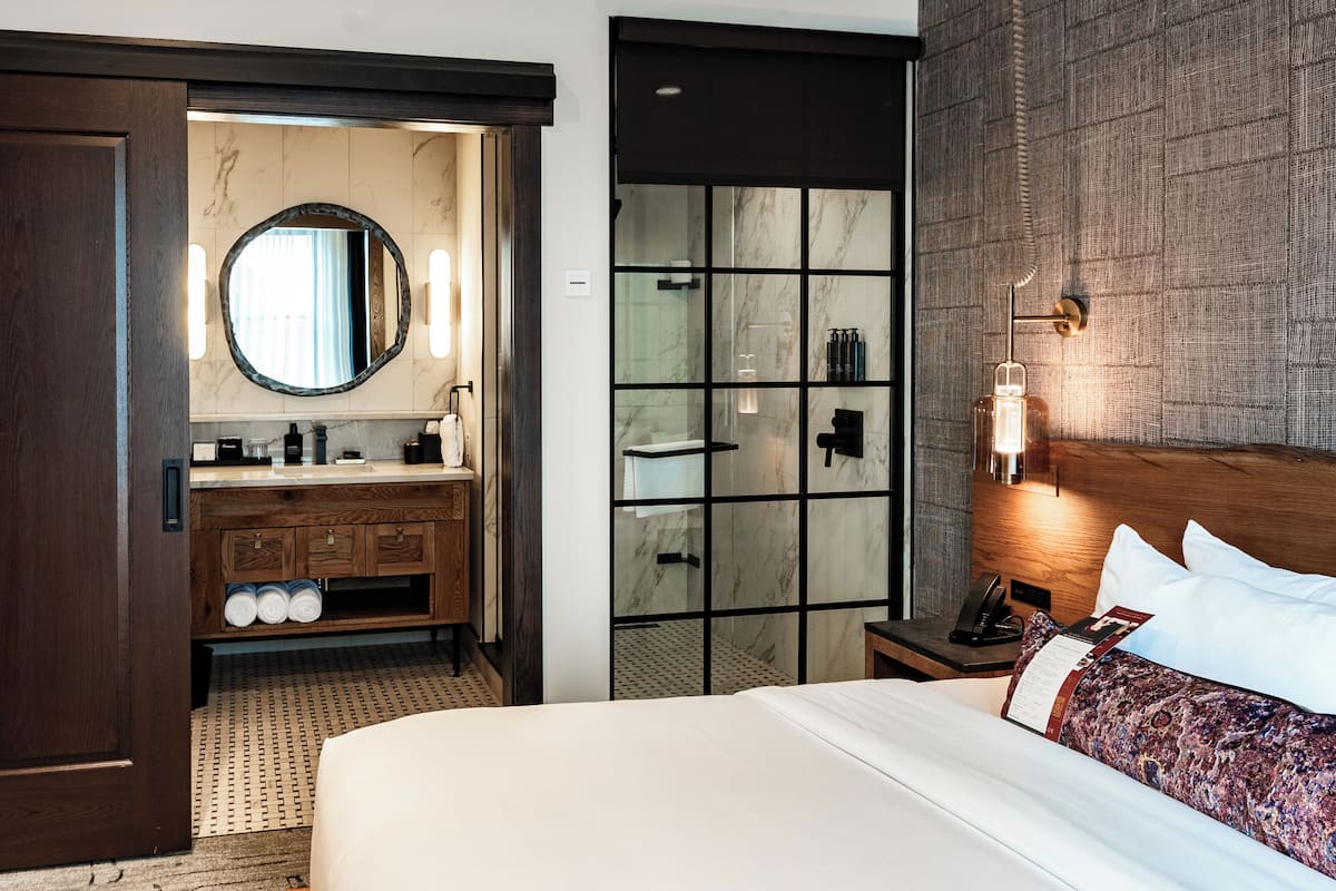 A hotel room with a large bed, a wooden vanity with a mirror, a built-in shower, and a nightstand with a lamp.
