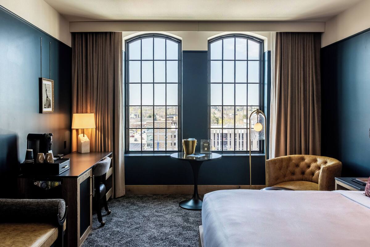 A hotel room with a bed, a desk with a lamp, a chair, and a view through large windows.