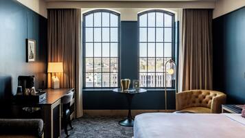A hotel room with a bed, a desk with a lamp, a chair, and a view through large windows.