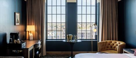 Ein Hotelzimmer mit einem Bett, einem Schreibtisch mit Lampe, einem Sessel und Blick durch große Fenster.