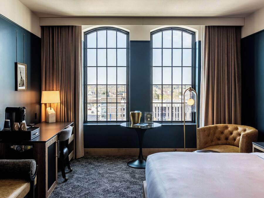 A hotel room with a bed, a desk with a lamp, a chair, and a view through large windows.