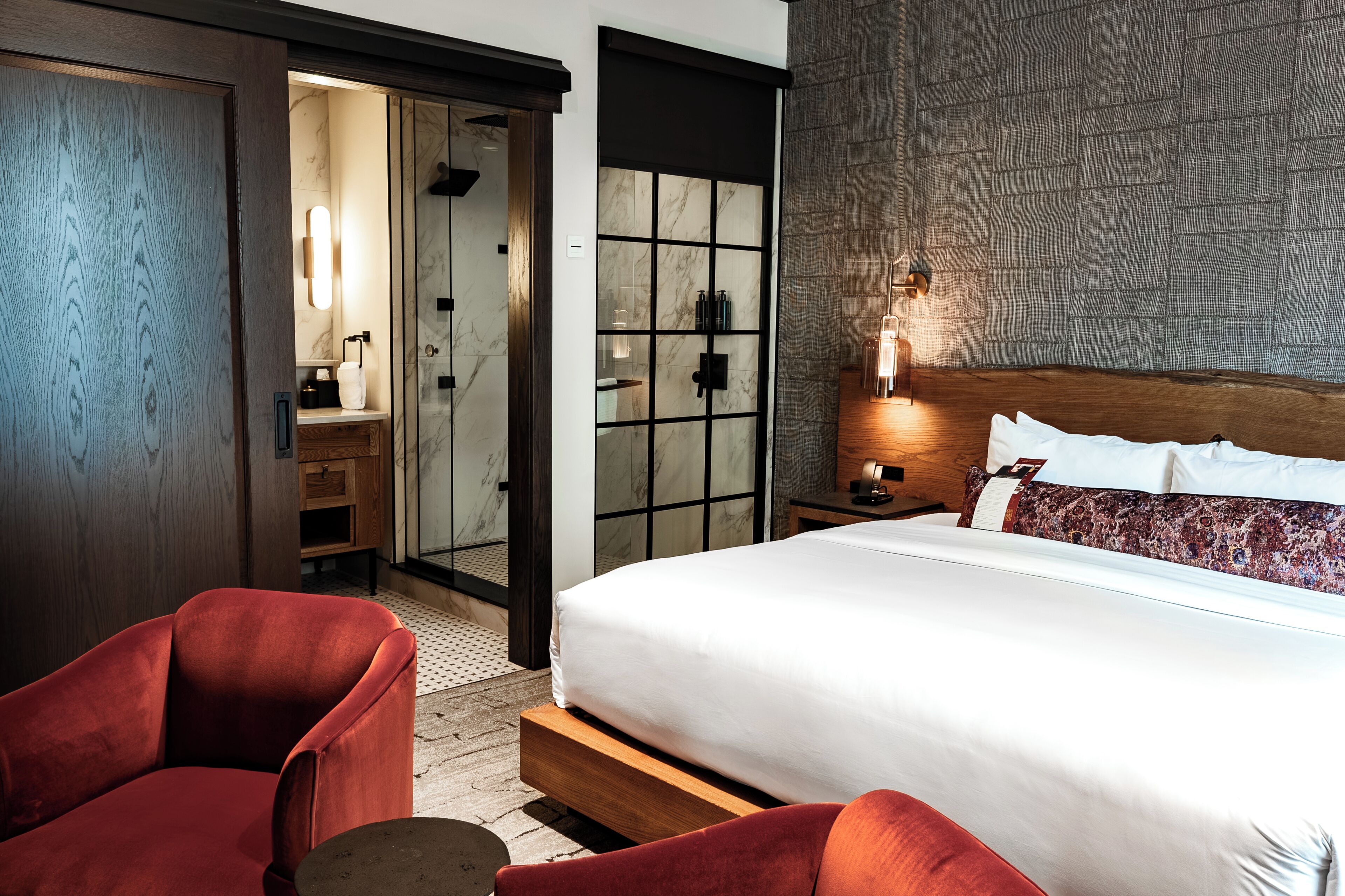 Une chambre d’hôtel moderne avec un grand lit, un fauteuil rouge, une table de chevet en bois, un miroir et une salle de bain avec douche et toilettes.