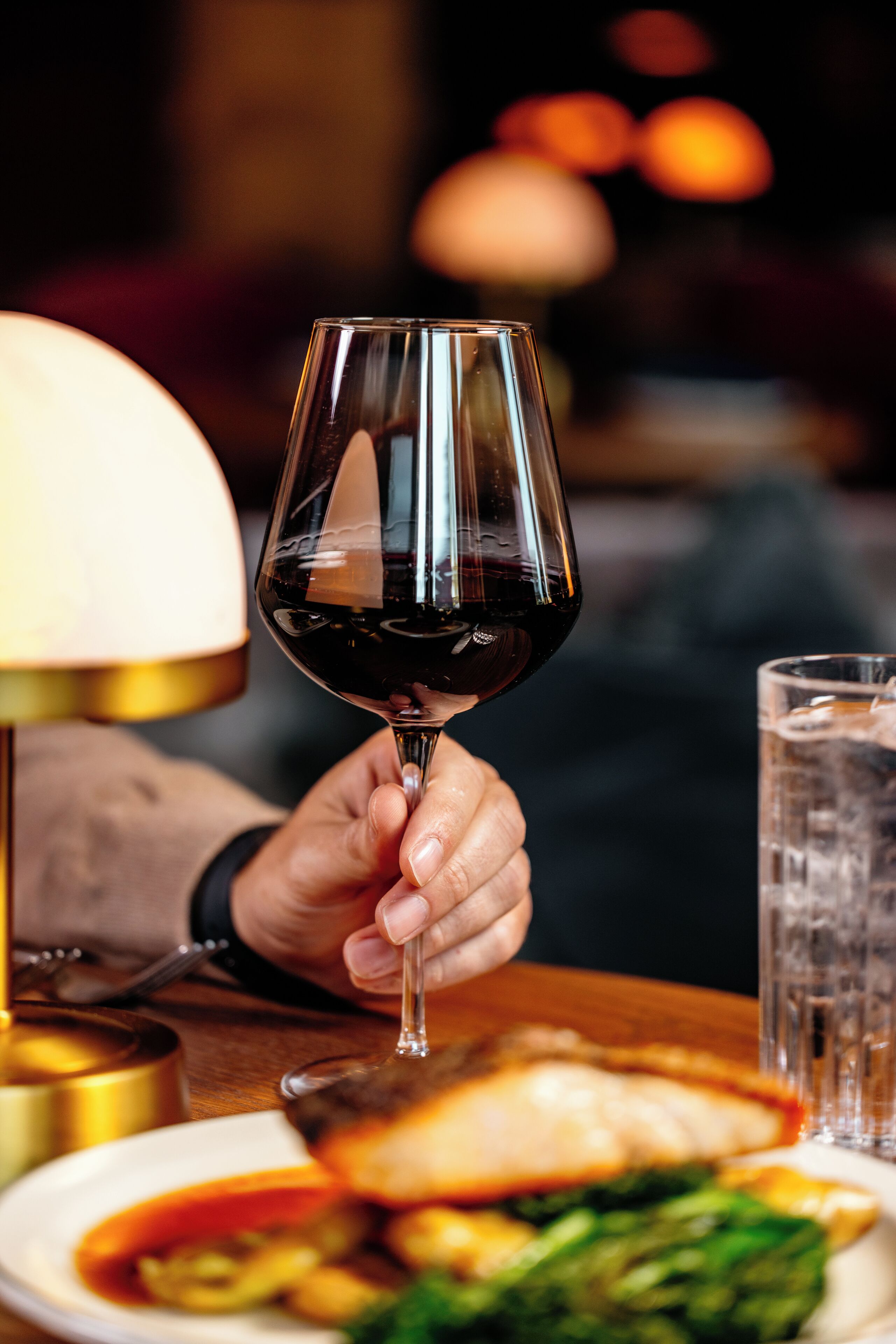 A hand holding a wine glass with a dark liquid, a plate with food, and a glass of water on a table.