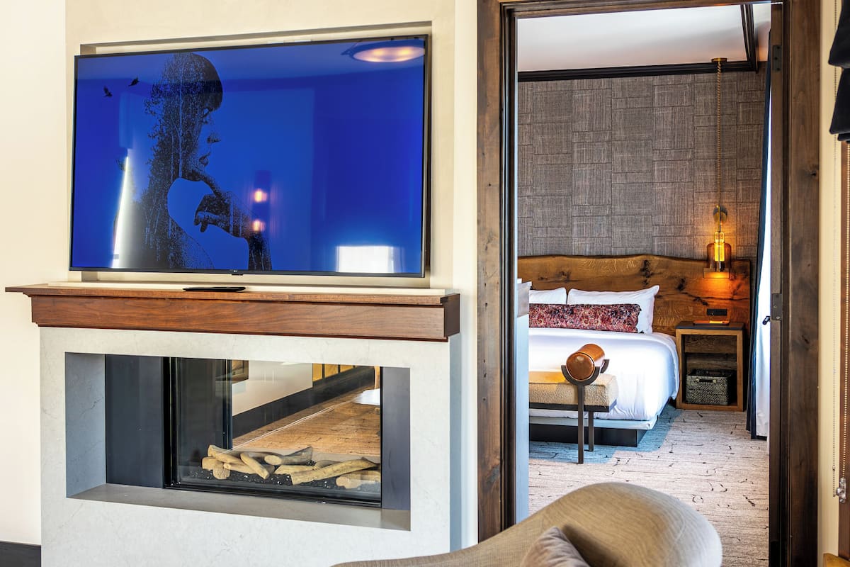 A modern hotel room with a television on a stand, a fireplace, a bed with a wooden headboard, and a nightstand with a lamp.