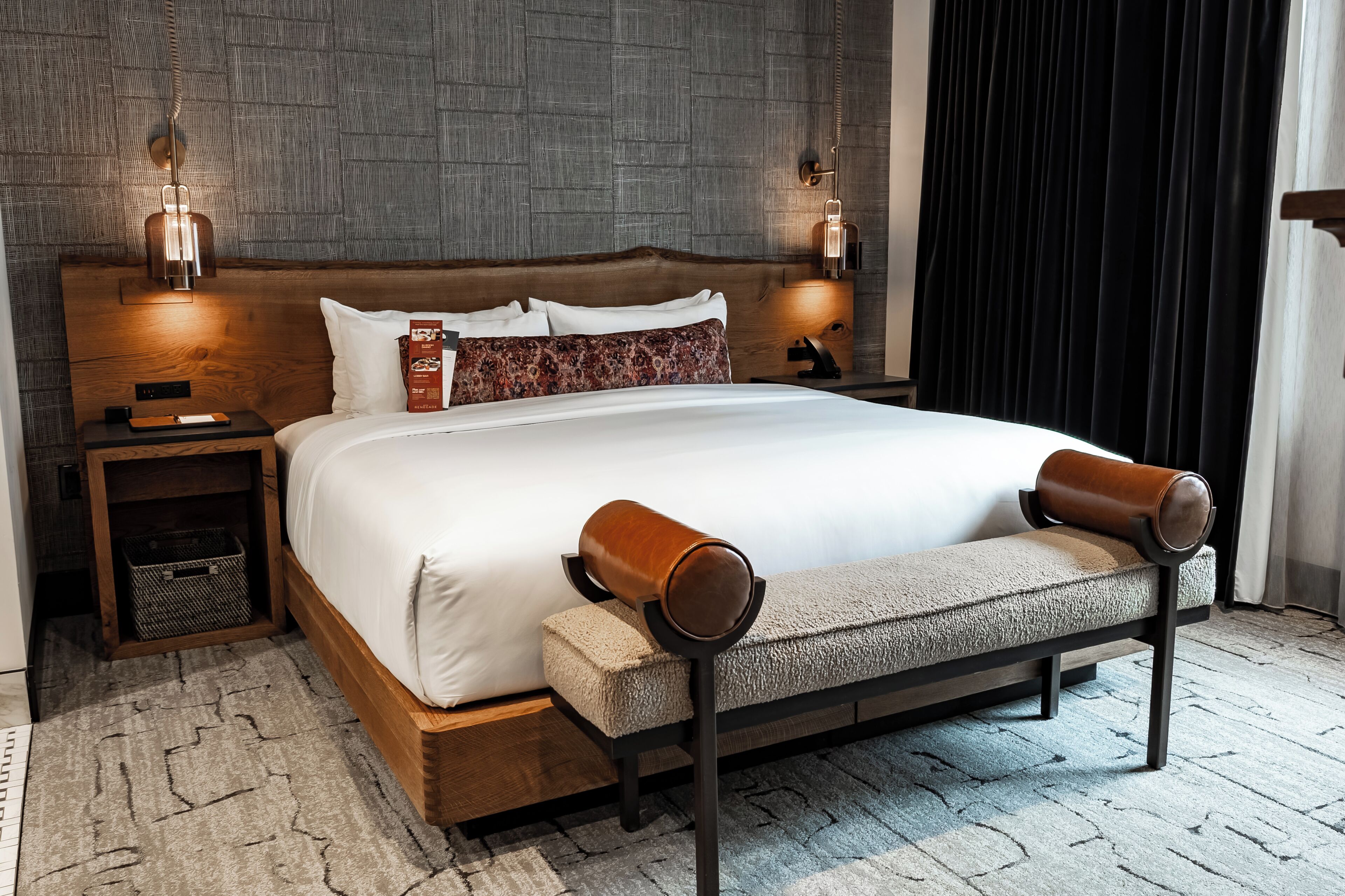A modern hotel room with a large bed, wooden headboard, two bedside lamps, a bench, and a textured rug.