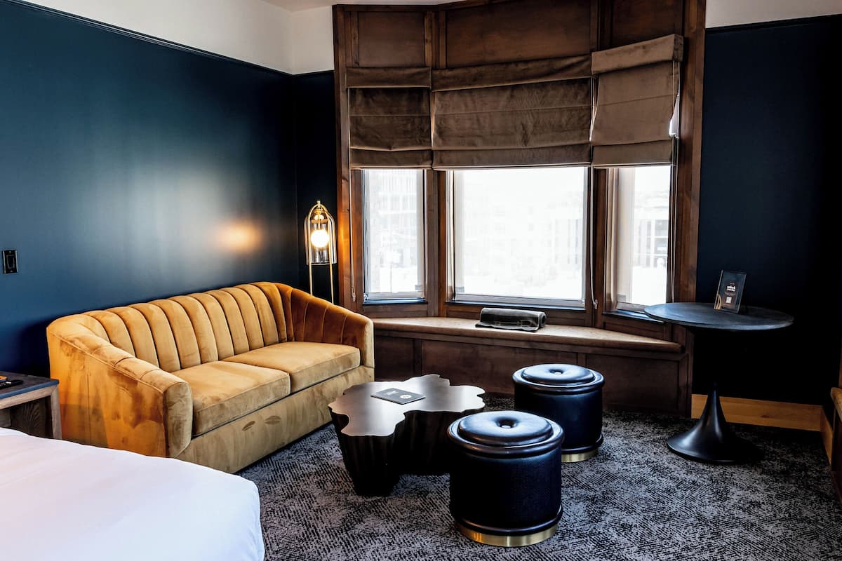 A hotel room with a sofa, a small table, two stools, and a window with blinds.