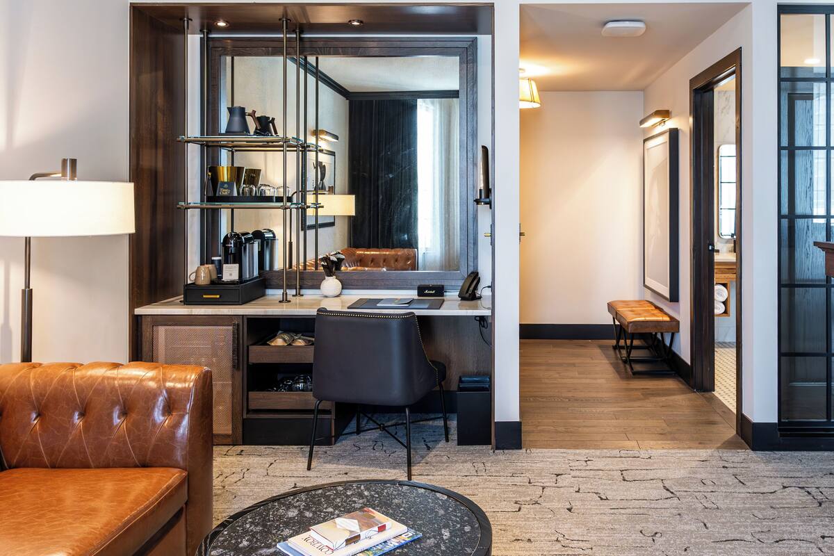 A modern hotel room with a leather sofa, a round coffee table, a desk with a computer, shelves with decorative items, and a large mirror.