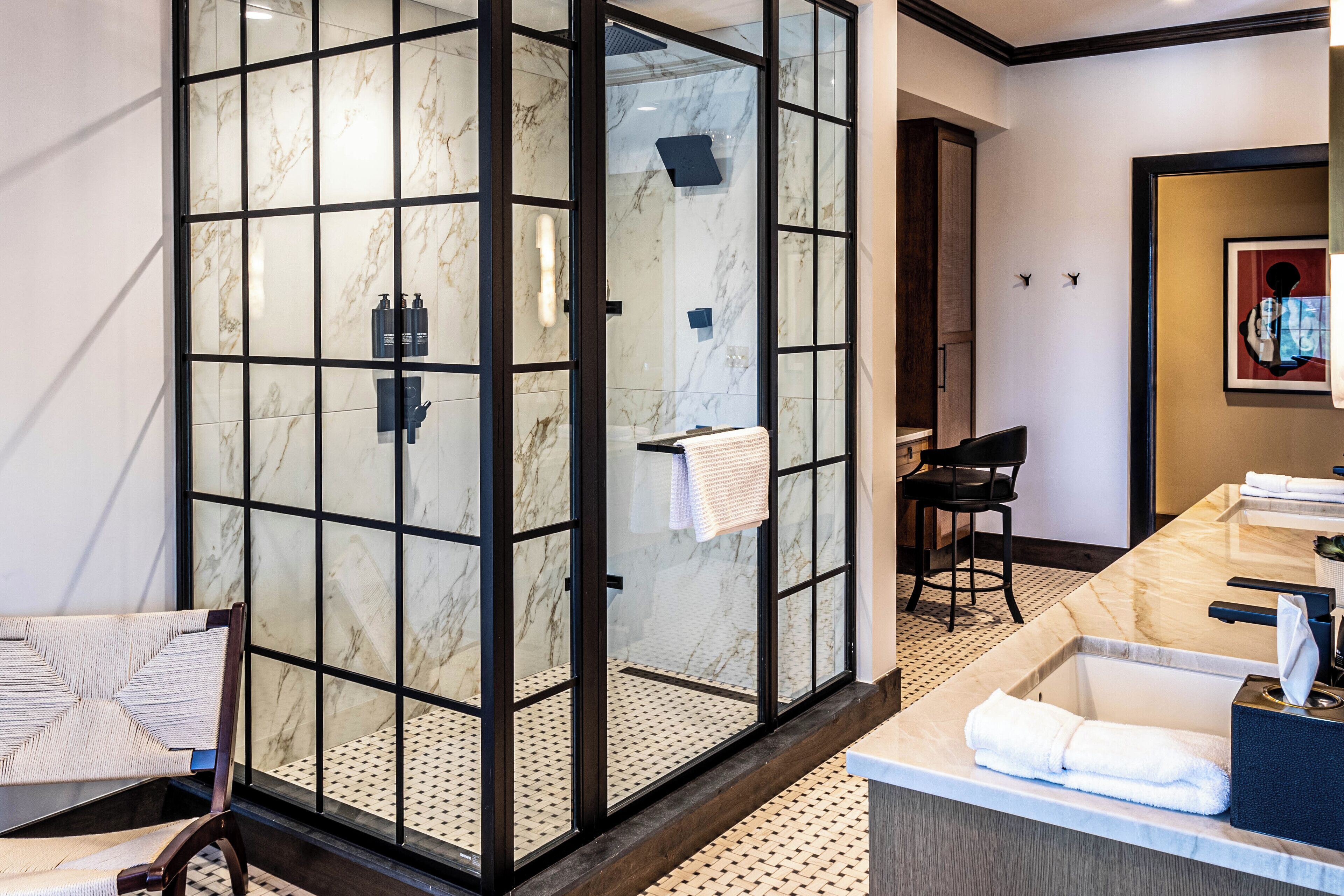 A modern bathroom with a glass-enclosed shower, a marble countertop, and a seating area with a chair.
