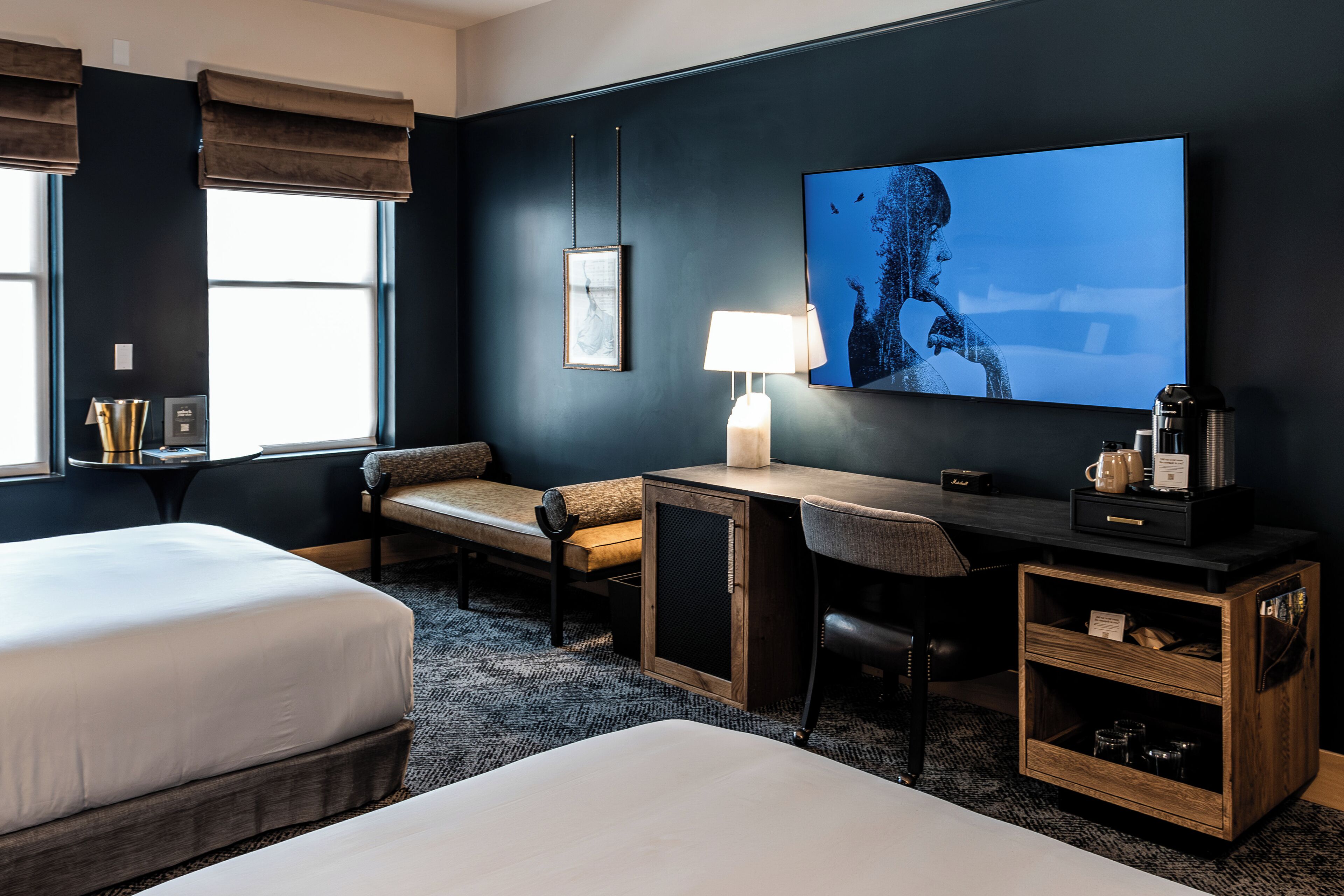 A hotel room with two beds, a desk, a chair, a lamp, a TV, and a coffee machine.