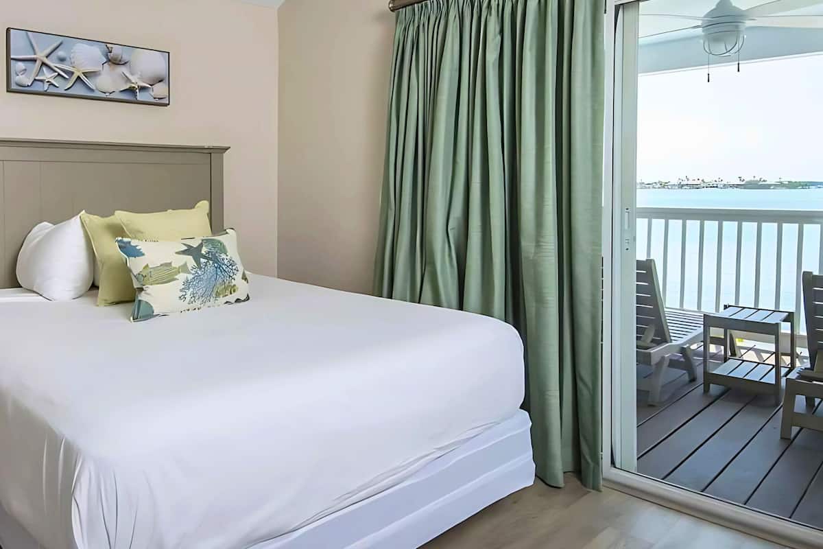 A hotel room with a bed, a nightstand, a lamp, a painting of seashells, a balcony with a view of the sea, and a table with chairs.