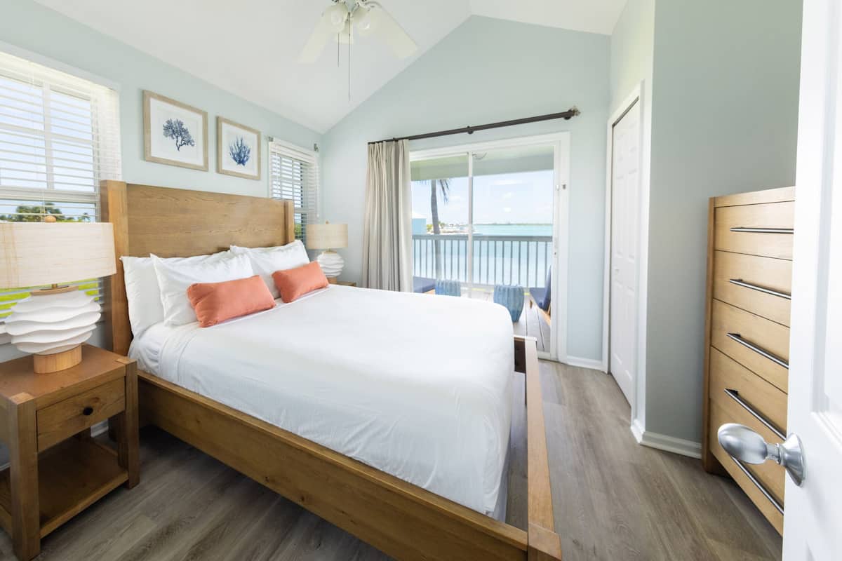 A bedroom with a wooden bed, white bedding, a nightstand with a lamp, and a view of the outdoors through a balcony door.