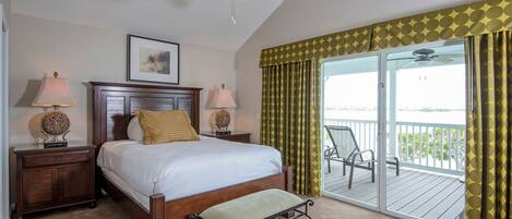 A hotel room with a bed, two bedside lamps, a wooden nightstand, a bench, a ceiling fan, a large window with a view, and a balcony with chairs.