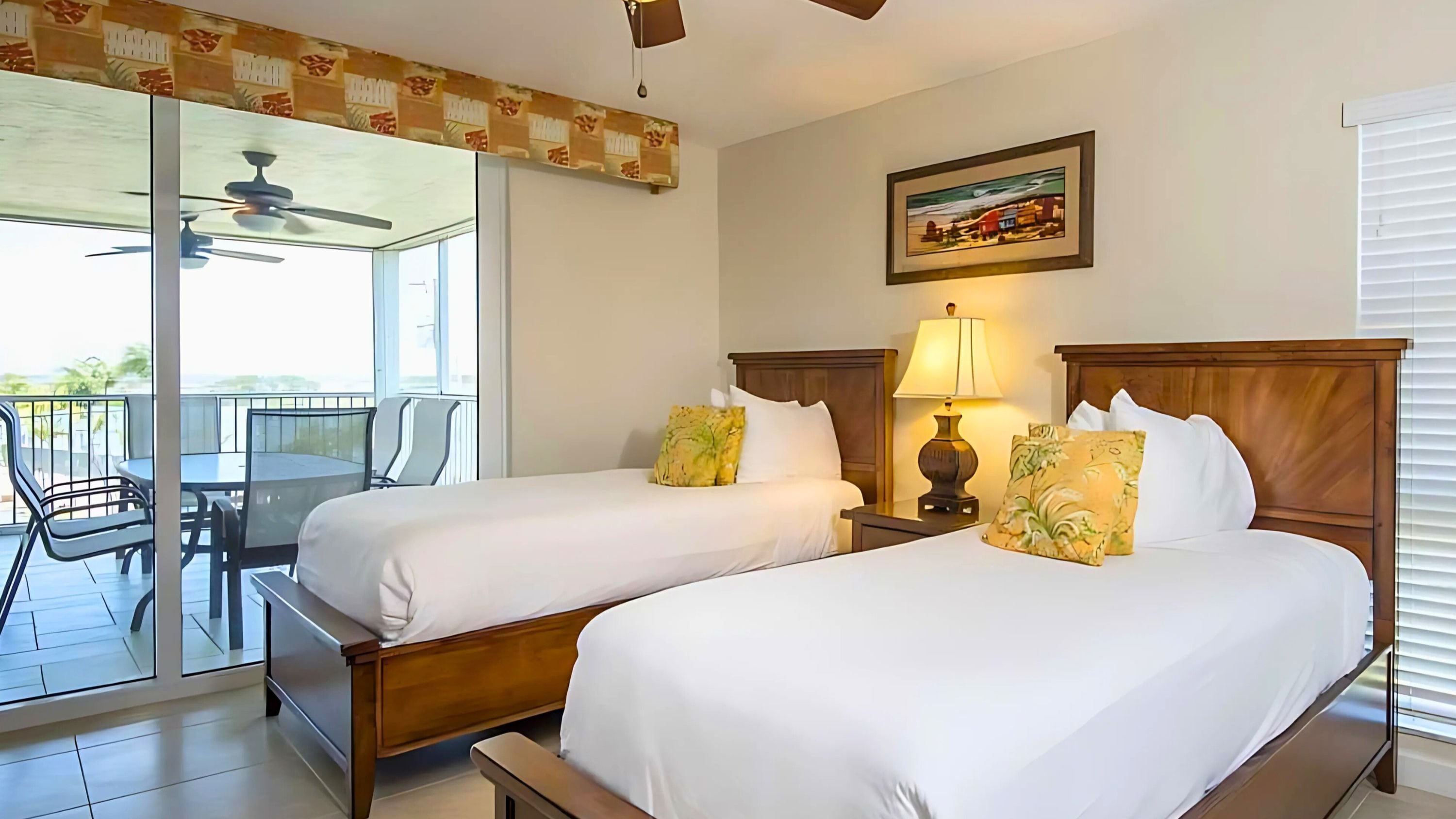 A hotel room with two beds, a balcony with a table and chairs, a ceiling fan, and a painting on the wall.