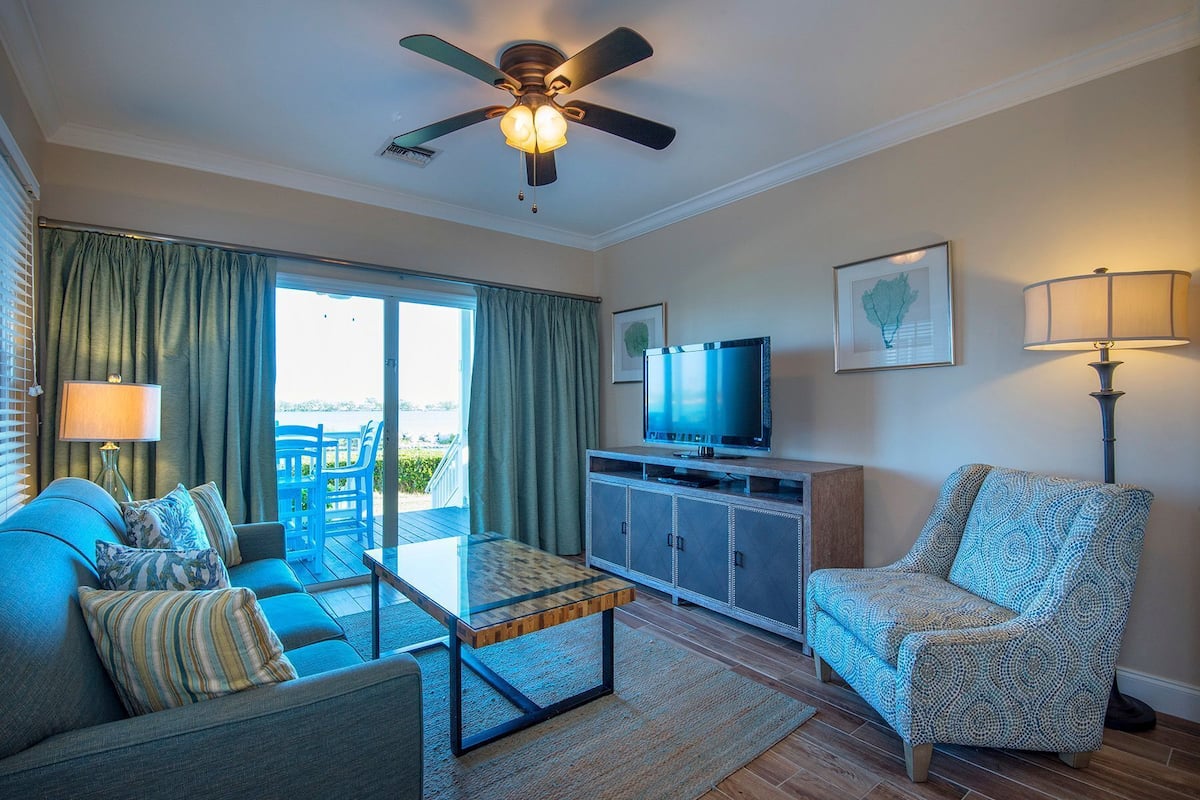 A living room with a sofa, armchair, glass coffee table, TV, and a ceiling fan.