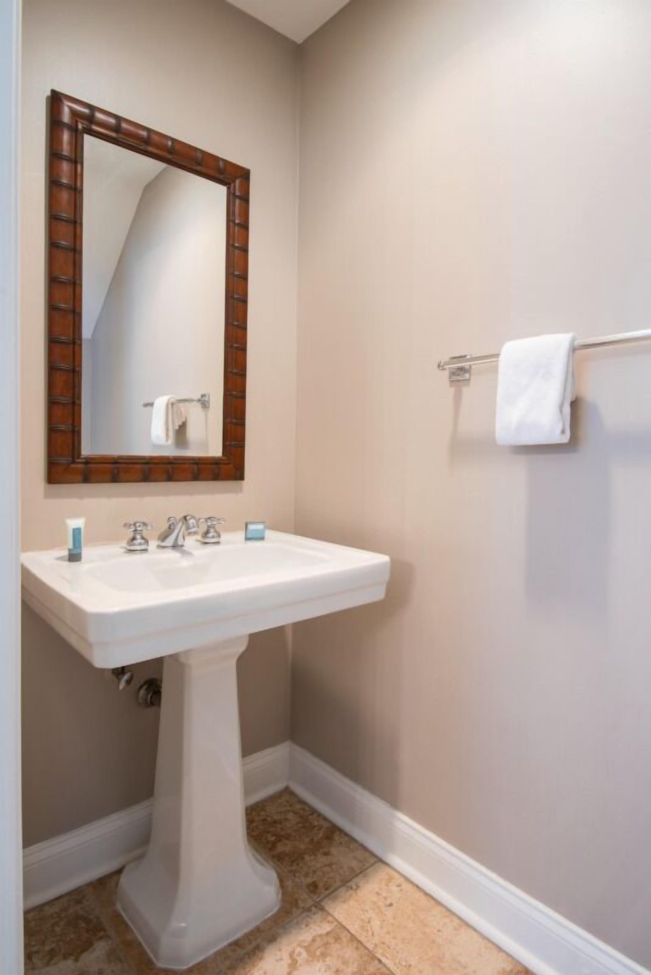 A bathroom with a pedestal sink, a wooden-framed mirror, and a towel rack.