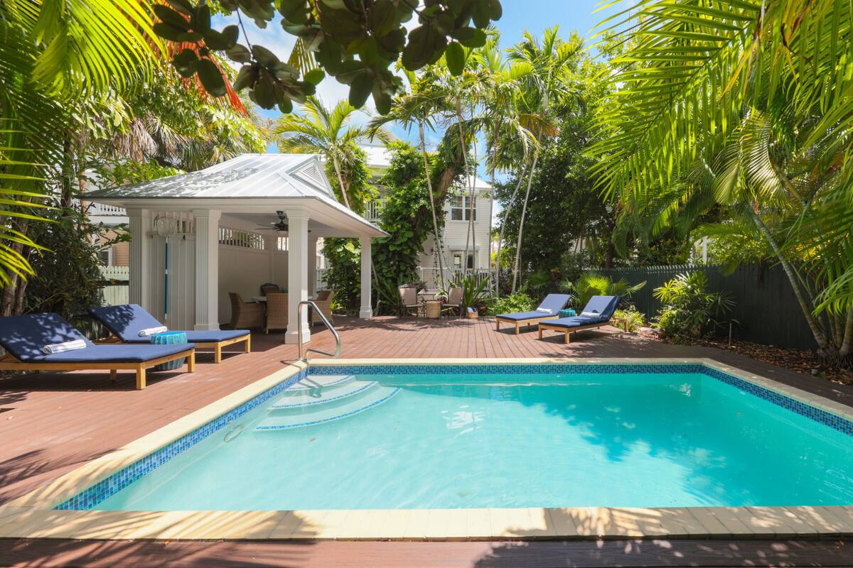 A swimming pool with lounge chairs, a covered seating area, and lush tropical vegetation.