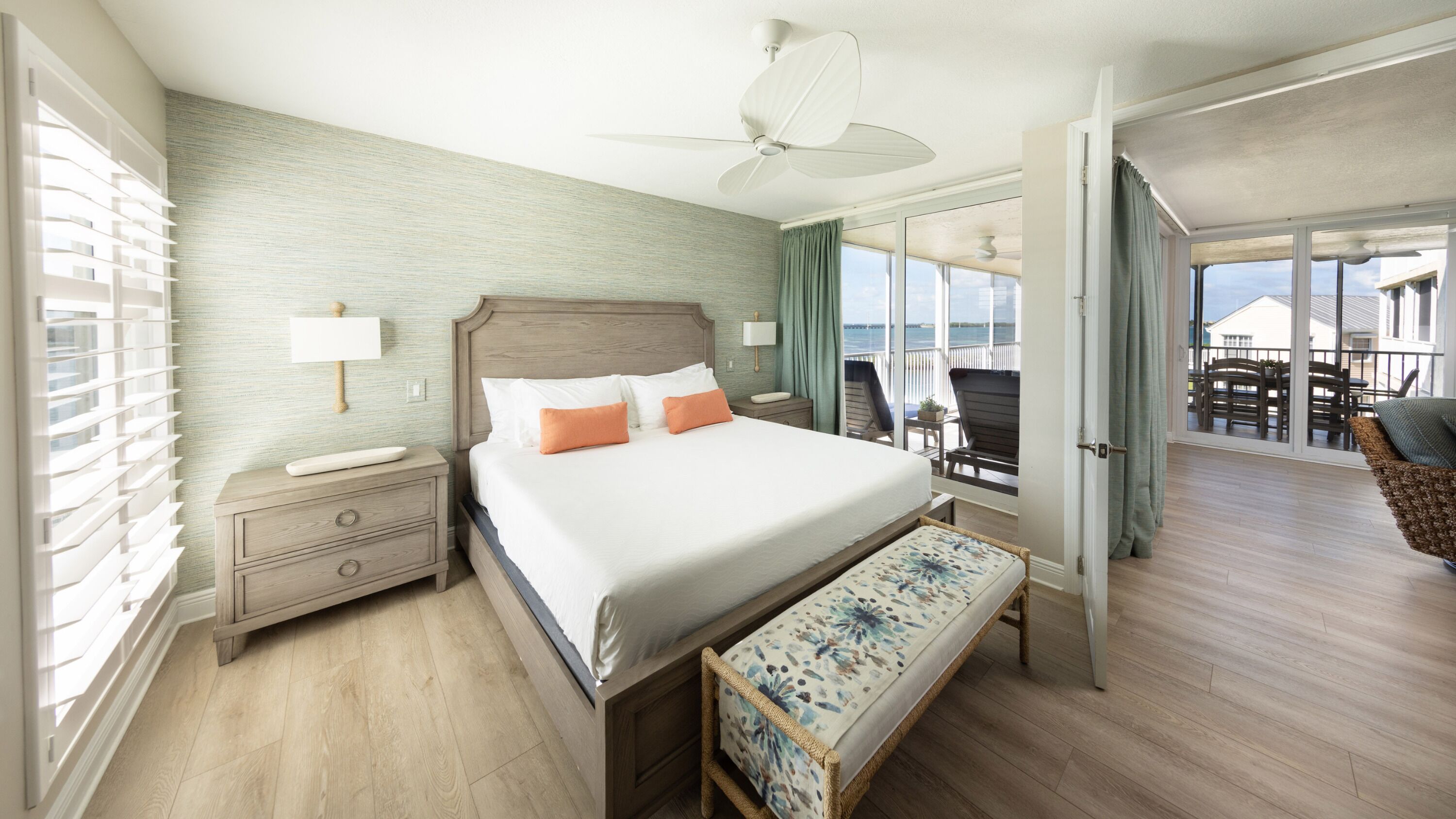 A spacious bedroom with a large bed, a bedside table, a nightstand, a bench, and a view of the balcony.