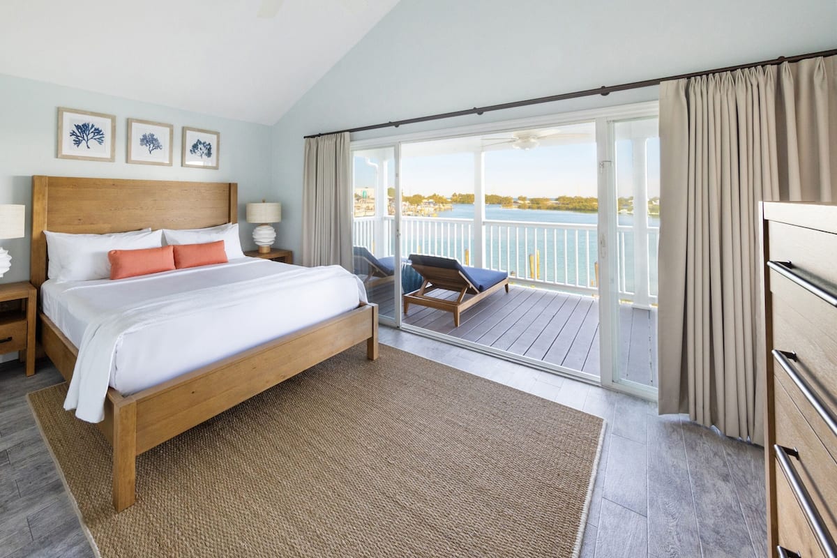 A bedroom with a wooden bed, a nightstand, a large window, and a view of the sea.
