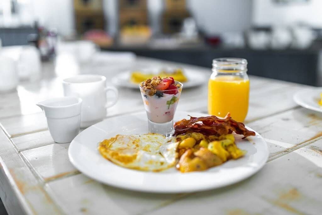 Une table dressée pour le petit-déjeuner avec des assiettes d’œufs, de bacon et de pain grillé, un verre de jus d’orange et un yaourt en verrine agrémenté de fruits et de granola.