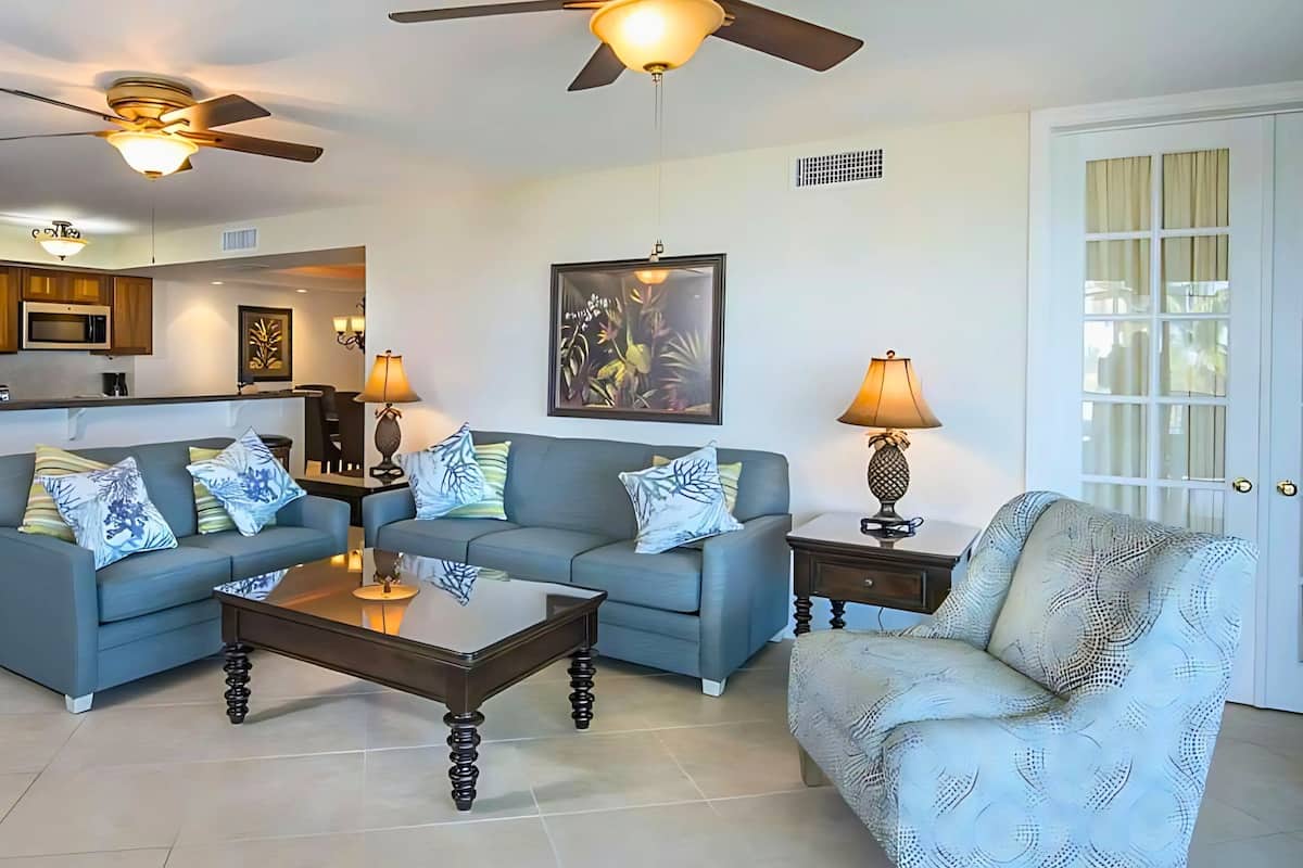 A living room with two sofas, a coffee table, and a patterned armchair. There are ceiling fans, a dining area, and a kitchen in the background.