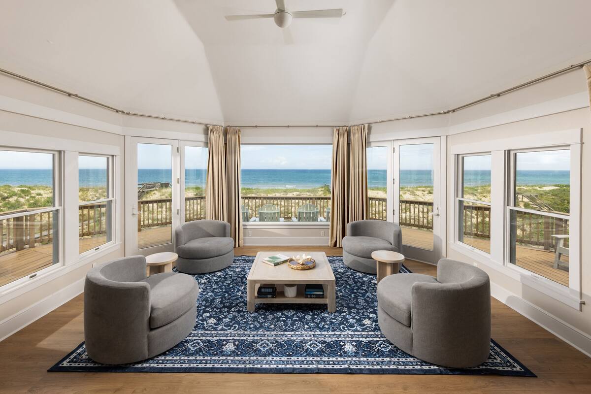 Habitación con vista a la playa, que incluye un ventilador de techo, dos sillones, una mesa de centro y una alfombra estampada.