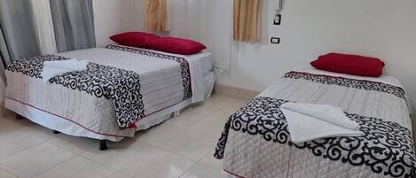 Two single beds with patterned bedding and red pillows in a room with a tiled floor and a window with curtains.