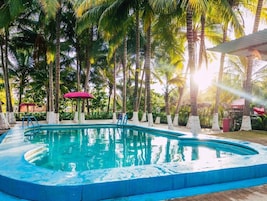 Piscina rodeada por palmeiras, com uma escada dando acesso à água e um guarda-sol vermelho por perto.