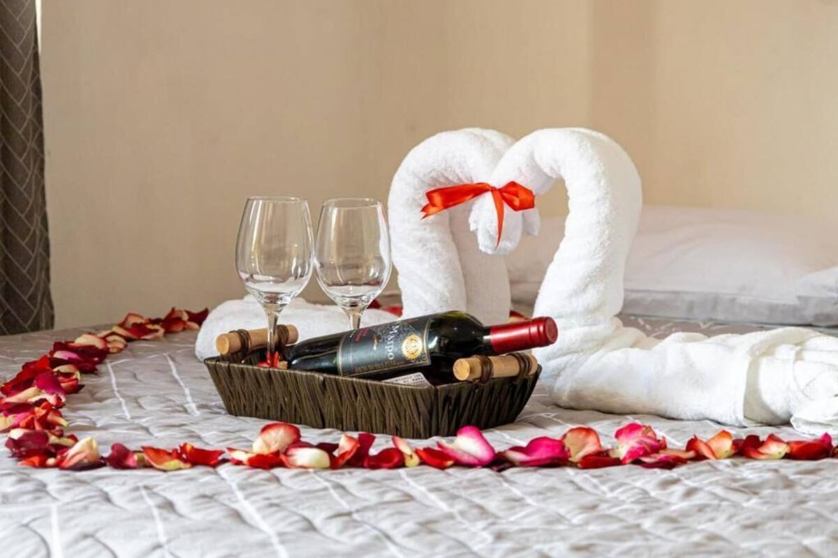 Una cama con una toalla dispuesta en forma de corazón, una botella de vino, dos copas y una cesta de pétalos de rosa.