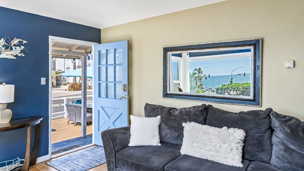 Un salon avec un canapé gris foncé, un coussin blanc et moelleux, un tableau encadré représentant une plage et une porte bleue donnant sur un espace extérieur.