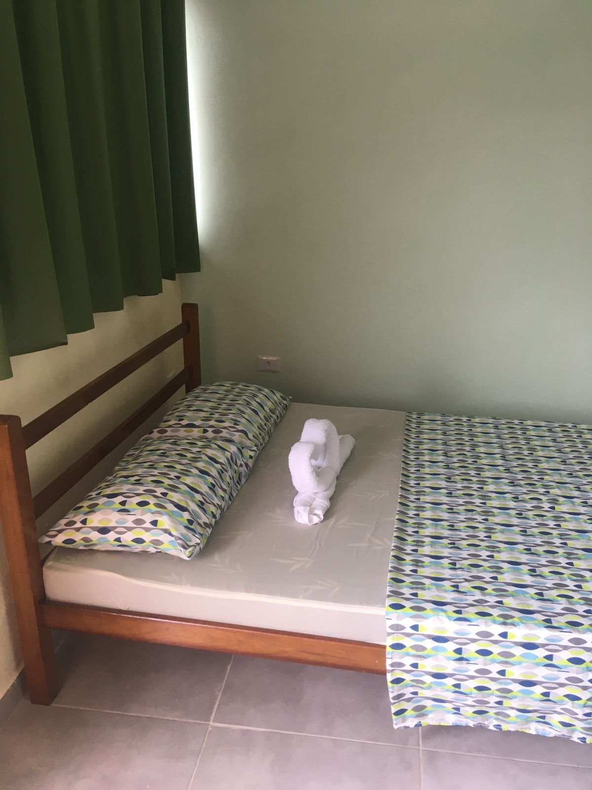 Uma cama de solteiro com colcha estampada e almofadas, cabeceira de madeira e cortina verde.