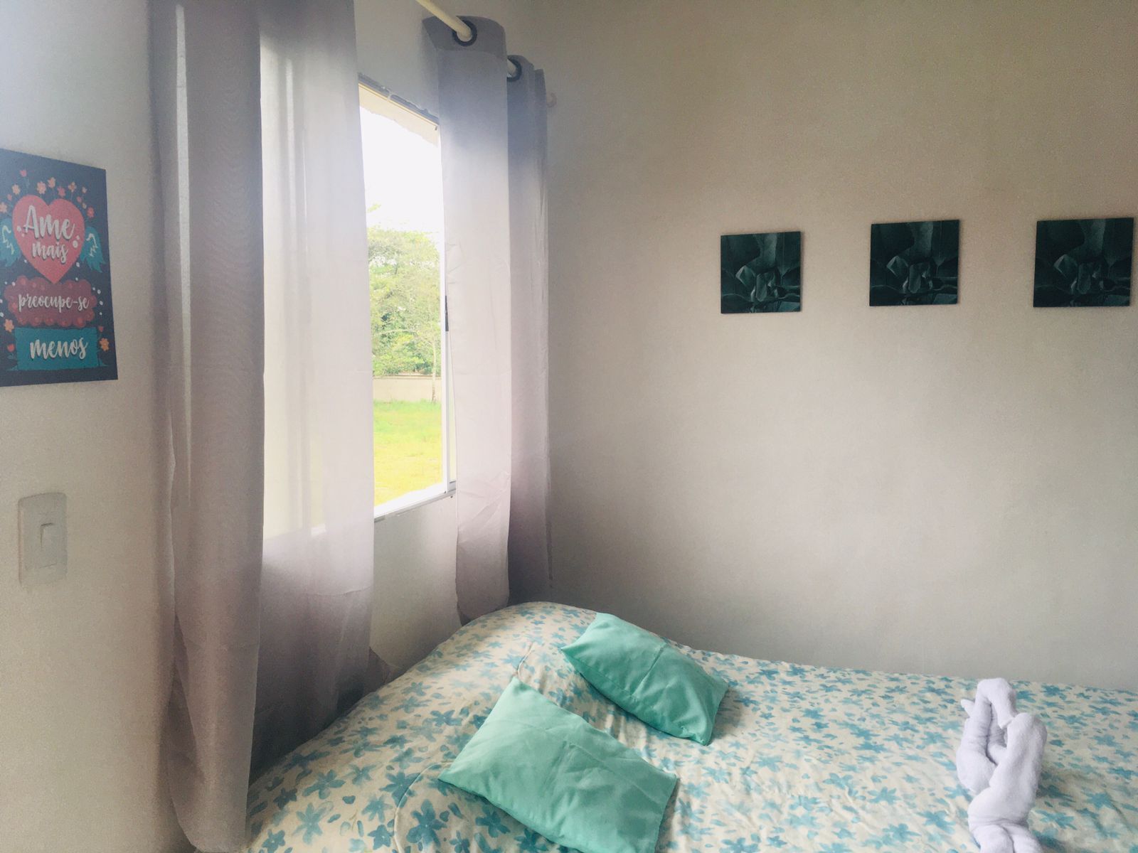 A bedroom with a bed, two pillows, a window with curtains, and three wall-mounted artworks.
