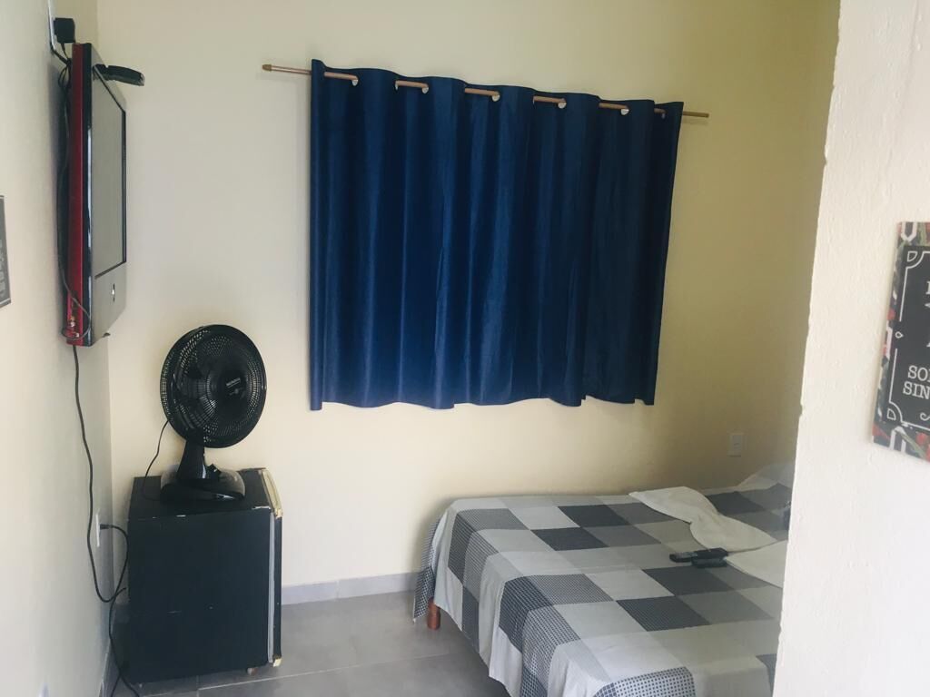 Een kleine kamer met een bed, een ventilator en een aan de muur bevestigde televisie.