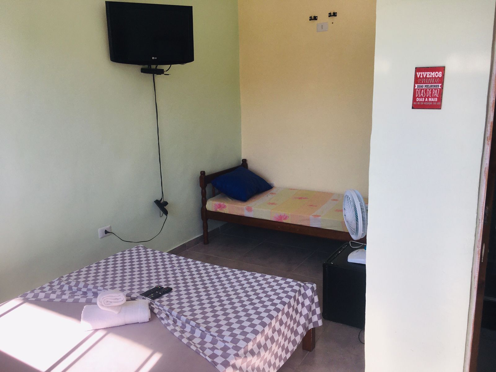 Una habitación pequeña y sencilla con una cama, una silla, un televisor montado en la pared y un ventilador en el piso.