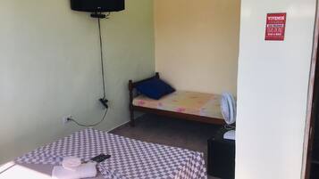 Una habitación pequeña y sencilla con una cama, una silla, un televisor montado en la pared y un ventilador en el piso.