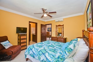 Quarto com uma cama, uma cadeira, televisão, ventoinha no teto e uma porta que dá para outro cômodo.