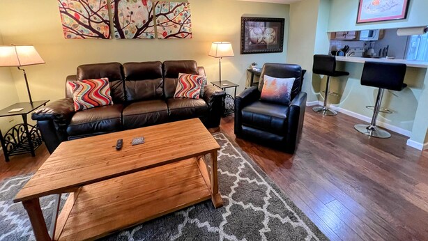 A living room with a brown leather sofa, a wooden coffee table, a black leather armchair, and a kitchen area with a bar stool. - Townhouse in the Pines
 (Bloomington)