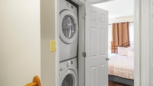 A laundry room with stacked washers and dryers, a door leading to a bedroom with a bed and curtains. - Townhouse in the Pines
 (Bloomington)