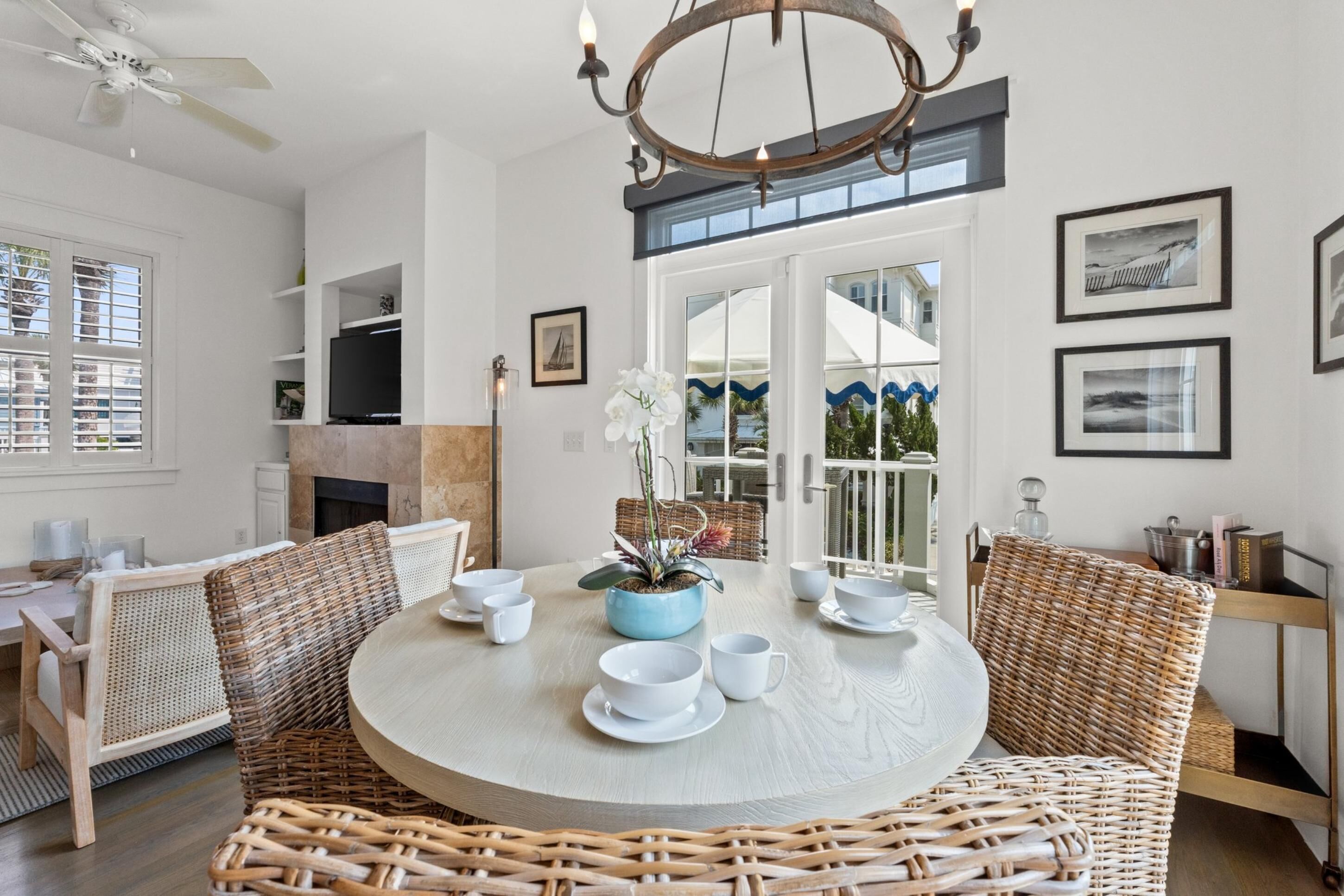 Una mesa redonda de comedor con platos y tazas blancos, rodeada de sillas de mimbre, en una habitación luminosa con paredes y ventilador de techo blancos.
