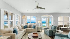 A spacious living room with a large window overlooking the ocean, a ceiling fan, and a cozy seating area with a coffee table. - Villas at Sunset Beach  (Panama City Beach)