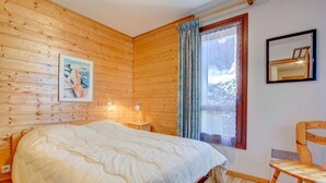 Een slaapkamer met houten lambrisering, een bed, nachtkastje, stoel, raam met gordijnen en een lijst met afbeelding.