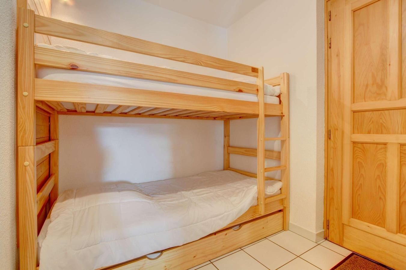 A bunk bed with a wooden frame and white bedding in a room with tiled flooring and a wooden door.
