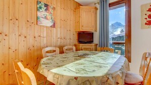 Een eethoek met een ronde tafel bedekt met een tafelkleed met olijfolieprinten, houten stoelen, een televisie op een houten kast en een raam met uitzicht op de bergen.