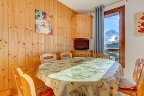 A dining area with a round table covered by a tablecloth with olive oil prints, wooden chairs, a television on a wooden cabinet, and a window with a mountain view. - 150m from resort center, 1st floor, balcony, tv, ski locker, 36m², Morzine (Morzine)