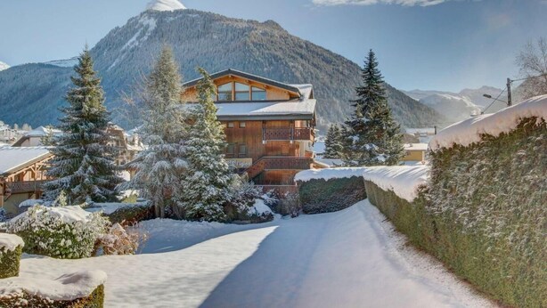 Een houten lodge met een balkon, omgeven door sneeuwbedekte bomen en bergen op de achtergrond.